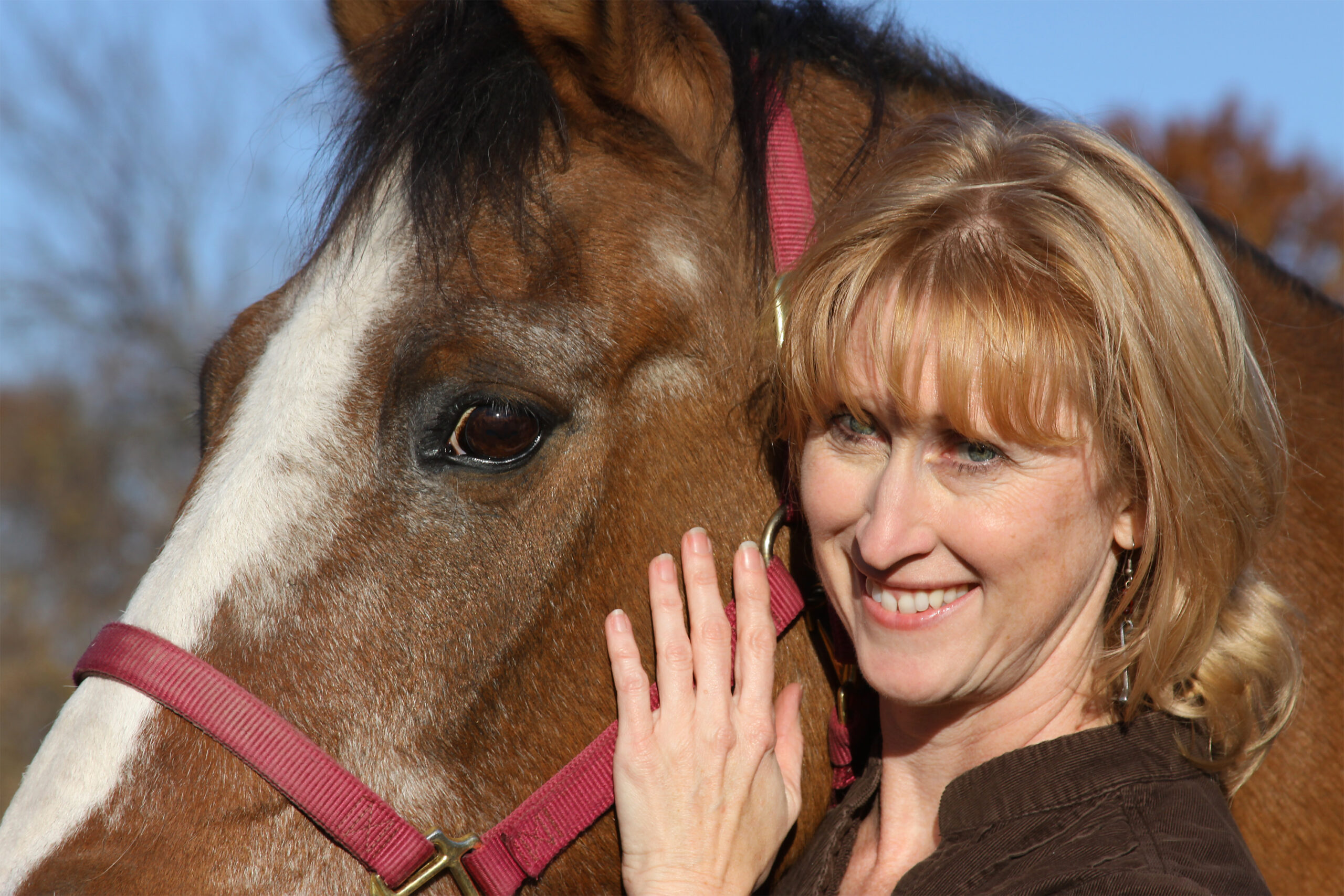 Jodi looks a little like Meryl Streep. She has red shoulder-length hair and hazel eyes. She is resting her head against the head of a horse with reddish brown hair and a white stripe down his nose.